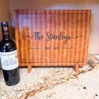 Personalized maple end grain family cutting board displayed on a wood stand beside a wine bottle