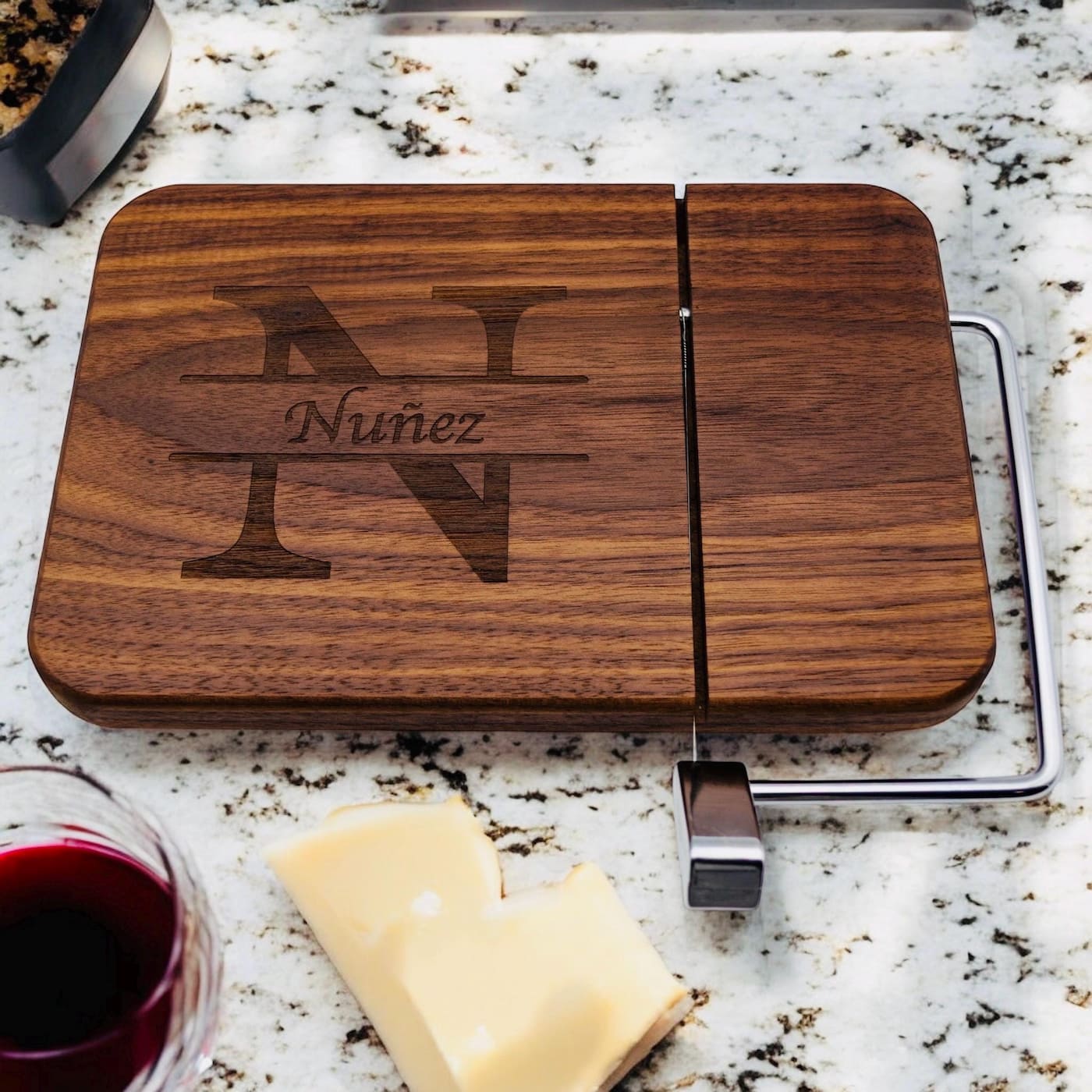 Personalized walnut cheese slicer with engraved split monogram beside wine glass and cheese on granite countertop
