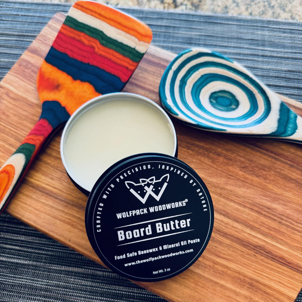 Open tin of Wolfpack Woodworks board butter on wooden cutting board with colorful kitchen utensils